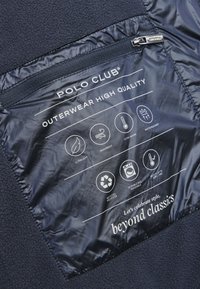 Marineblå fleecjacka med en vattentät, zip-försluten ficka märkt med skötselråd och prestandaikoner för hållbarhet och värme.