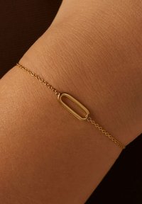 Bracelet en chaîne dorée avec un maillon ovale allongé, texture lisse et fine chaîne, porté sur un poignet avec une texture de peau visible.