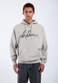 Sudadera beige con gráfica negra de firma, capucha con cordón, bolsillo tipo canguro y puños acanalados; combinada con jeans gris oscuro.