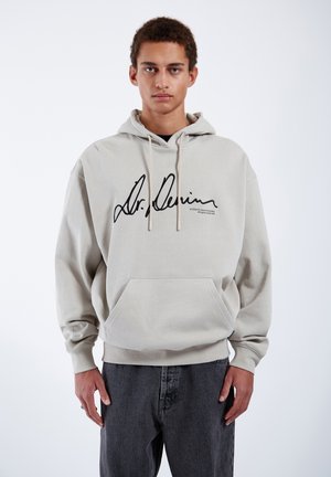 Beige hoodie with black signature graphic, drawstring hood, kangaroo pocket, and ribbed cuffs; paired with dark gray jeans.