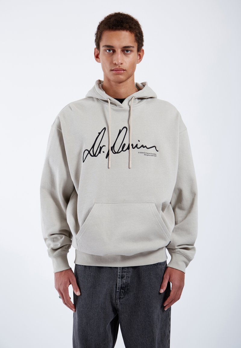 Sudadera beige con gráfica negra de firma, capucha con cordón, bolsillo tipo canguro y puños acanalados; combinada con jeans gris oscuro.