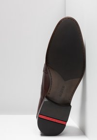 Chaussure en cuir marron avec un bout pointu et des détails cousus. La semelle est noire avec une bande accent rouge et un logo de marque sur le talon.