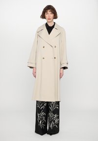 Beige double-breasted trench coat with large lapels and gold-tone buttons, paired with black wide-leg pants featuring white floral patterns.