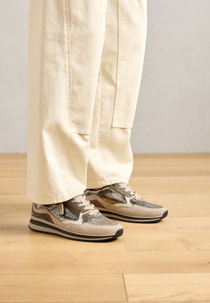 Μπεζ και καφέ υφαντά sneakers που φοριούνται με χαλαρούς κρεμ παντελόνια, σε μια ανοιχτόχρωμη ξύλινη επιφάνεια.