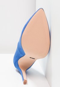 Chaussure à talon stiletto en daim bleu à bout pointu, vue de la semelle vers le haut sur fond blanc.