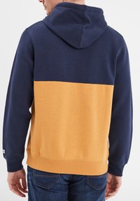 Sudadera con capucha con parte superior azul marino y parte inferior mostaza. Textura suave, puños acanalados y detalle del logo en el dobladillo inferior.