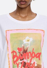 Camiseta blanca con una mano sosteniendo una flor, con un gráfico colorido que dice "Bimba y Lola" y un corazón. Material de algodón suave, con cuello redondo.