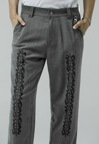 Personne portant un pantalon à carreaux gris avec des motifs d'écriture abstraite noire verticale sur les deux jambes, les mains dans les poches et une chaîne argentée du côté droit.