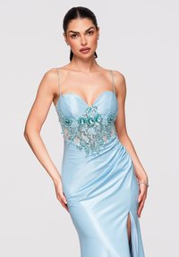 Femme aux cheveux foncés en queue de cheval lisse, portant une robe en satin bleu clair ornée de perles florales sur un corsage transparent et une fente haute jusqu’à la cuisse.