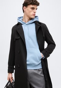 Νεαρός άνδρας που φοράει μαύρο παλτό τύπου τρεντσόρ πάνω από ανοιχτό μπλε φούτερ με κουκούλα και γκρι παντελόνι, κρατώντας μαύρη τσάντα, κοιτάζει προς τα πλάγια μπροστά σε λευκό φόντο.