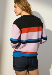 Pull en tricot multicolore avec des rayures noires, roses, rouges, bleues et oranges. Porté avec un short en jean, affichant une coupe décontractée.