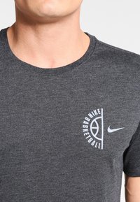 Mężczyzna w ciemnoszarej koszulce Nike do koszykówki z białym logo oraz grafiką boiska do koszykówki na klatce piersiowej.