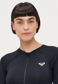 Chemise de surf sombre en tissu extensible, avec une fermeture éclair et un petit logo blanc en haut à gauche. Texture lisse, design près du corps.