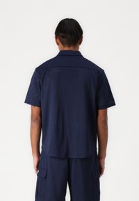 Marineblauwe korte mouwen shirt met geperforeerde textuur, voorzien van een klassieke kraag en een ontspannen silhouet. De achterkant laat subtiele ontwerpelementen zien.