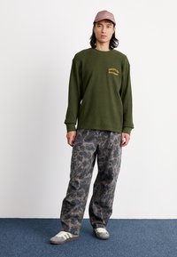 Olivza zaļas krāsas garām piedurknēm krekls ar dzeltenu uzrakstu, saskaņots ar pelēcīgiem kamuflāžas kravas biksēm. Brūna cepure un pelēki sporta apavi noslēdz apģērba komplektu.