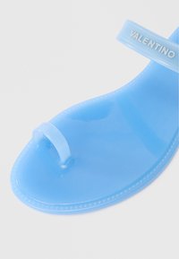 Kék gumipapucs áttetsző pánttal és domború logóval. Enyhén fényes, sima felületű, lekerekített orrú.