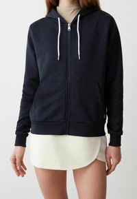Marinsblå zip-up hoodie med en frontficka, vita dragsnören och ribbade ärmslut. Buren över en krämfärgad kjol. Slät tygtextur.