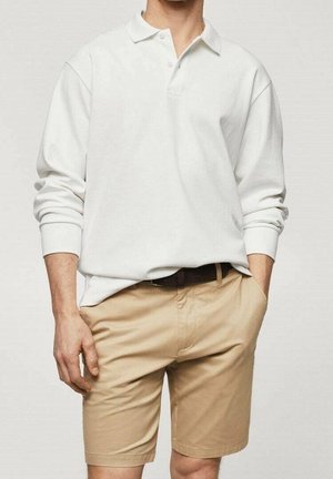 Biała koszulka polo z długim rękawem, kołnierzykiem i pojedynczą plisą na guziki, w połączeniu z jasnobeżowymi szortami i ciemnym paskiem.