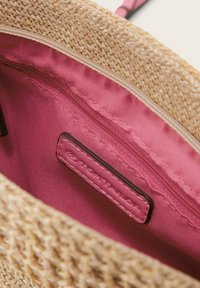 Vnitřek tkané slaměné tašky s hladkým růžovým textilním podšívem, zipovou kapsou a malou značkovou nálepkou z hnědé kůže.