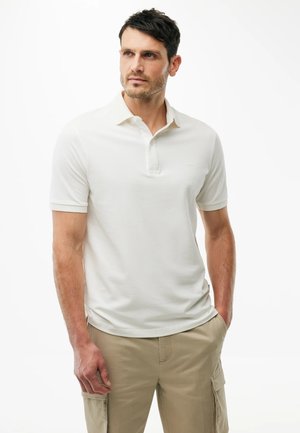 Mężczyzna ubrany w biały, krótki rękaw polo i beżowe spodnie typu cargo, pozujący z jedną ręką w kieszeni na białym tle.