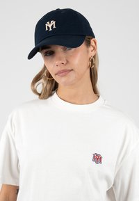 Joven mujer con gorra azul marino y camiseta blanca con logo bordado a juego, y pendientes de aro dorados, sobre un fondo gris.