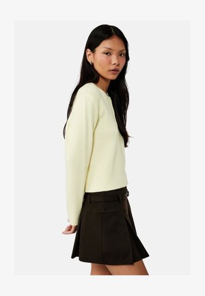 Mujer con cabello largo y oscuro, vistiendo un suéter crema acanalado y una minifalda negra plisada, de pie de perfil sobre un fondo liso.