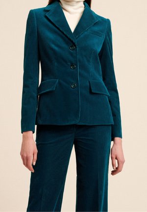 Femme portant un blazer en velours côtelé bleu sarcelle avec trois boutons et un pantalon large assorti, par-dessus un pull à col roulé clair.