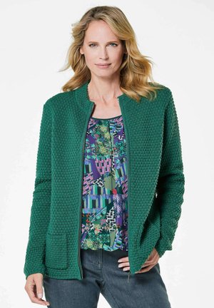 Veste zippée verte texturée avec un col rond, associée à un haut à motif coloré et à un jean foncé. Détails notables des poches sur la veste.