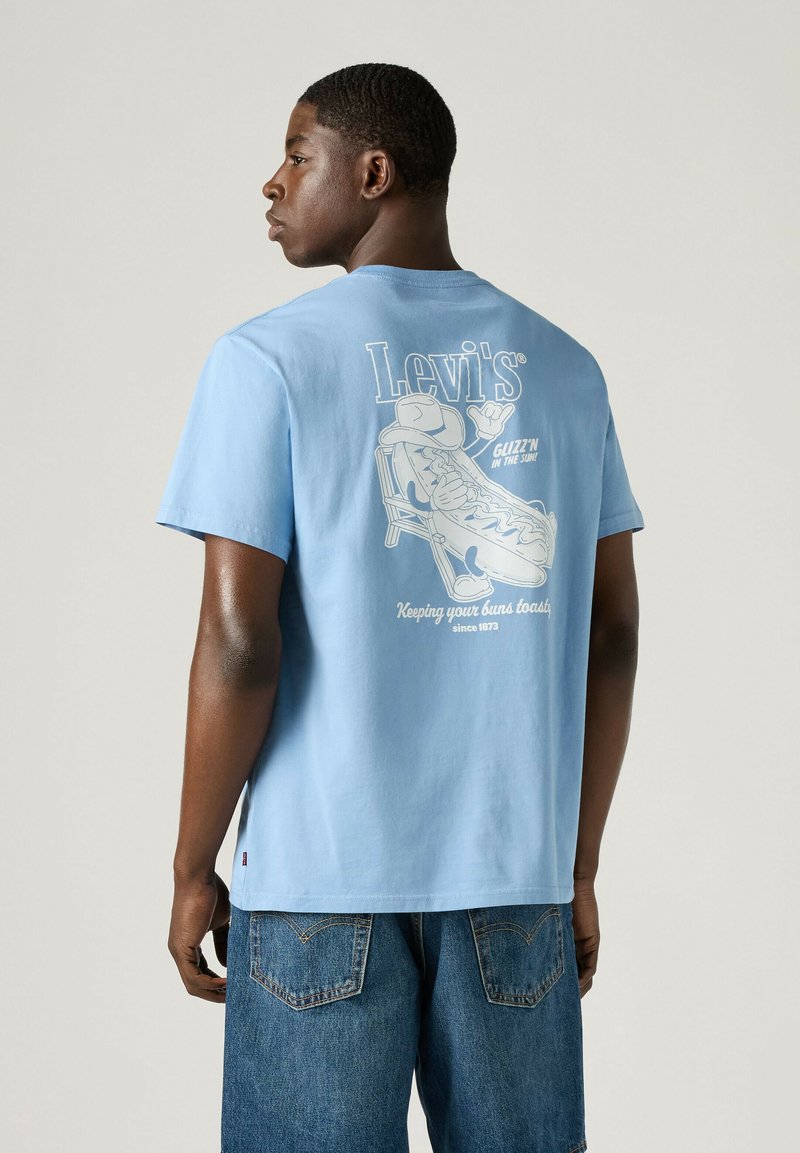 Junger Mann trägt ein hellblaues Levi's T-Shirt mit einer Grafik eines Hotdogs, der auf einem Stuhl liegt, und dem Text "Keeping your buns toasty" auf dem Rücken.