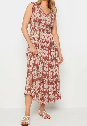 Kobieta w bezrękawnikowej, rdzawoczerwonej sukience midi z białymi kwiatowymi wzorami i rozcięciem po boku, w połączeniu z białymi, płaskimi sandałami na paski.
