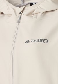 Beżowa kurtka z kapturem wykonana z lekkiego materiału. Posiada pełny zamek błyskawiczny, teksturowaną powierzchnię oraz szare logo "TERREX" z przodu.