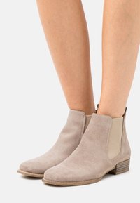 Zamszowe botki za kostkę w jasnobeżowym kolorze z elastycznymi panelami po bokach i niskim, warstwowym obcasem. Teksturowane wykończenie i zaokrąglony kształt noska.