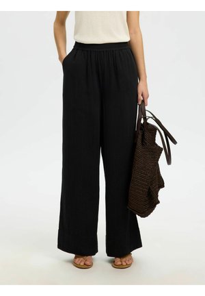 Donna che indossa pantaloni neri a gamba larga, top beige senza maniche, sandali color cuoio, con una borsa tote intrecciata marrone al fianco.