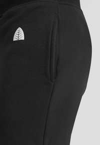Black sweatpants featuring a side pocket and a small leaf logo. The fabric appears soft and textured, suitable for casual wear.