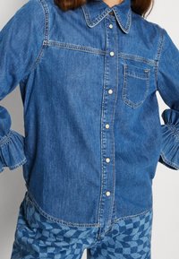 Ljusblå denimskjorta med krage, knapplåst och ficka. Har elastiska ärmslut, mjuk textur och kontrasterande sömdetaljer.