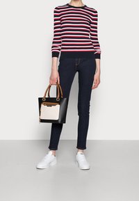 Black and cream handbag with brown handles, rounded shape, and a front pocket. Model wears a red and white striped sweater, dark jeans, and white sneakers.