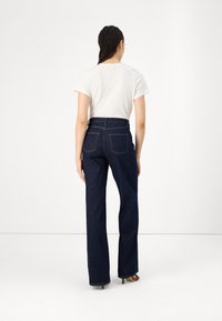 Dunkelblaue Hochbundjeans aus Denim mit geradem Beinschnitt, zwei aufgesetzten Taschen hinten und genähten Details. Tragen mit einem weißen Hemd.