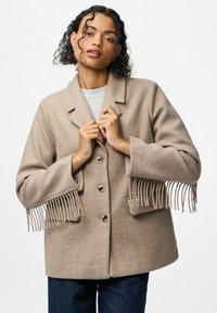 Beež villane mantel, millel on kohandatud disain, notšatud krae, ees nupud ja sulturid detailid käsivarte ääres.