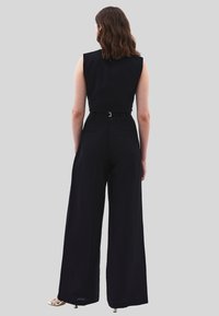 Zwarte mouwloze jumpsuit met hoge taille en brede benen. Heeft een ceinture-detail bij de taille en subtiele achterzakken. Soepele textuur.