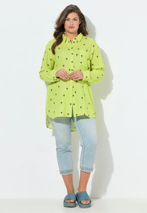 Frau trägt ein langes limettengrünes Hemd mit schwarzen Palmenmotiven, hellblaue kurze Jeans und blaue offene Sandalen.