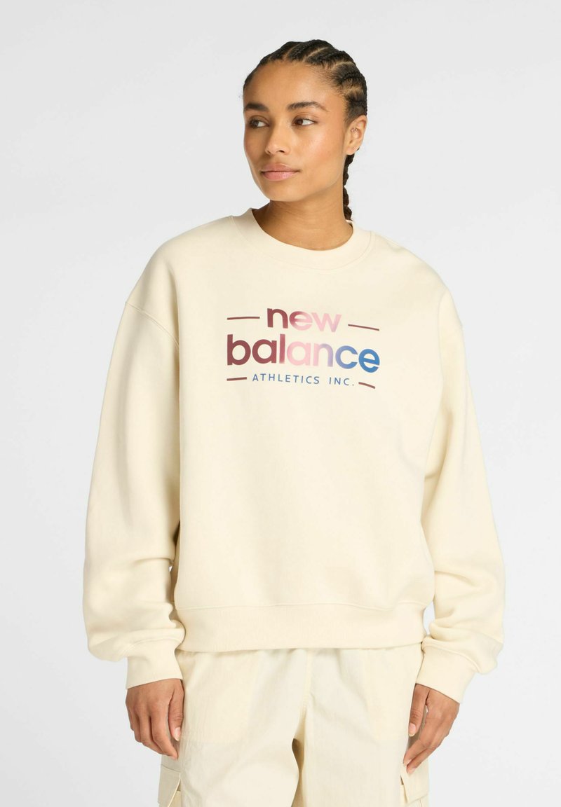Sweatshirt de couleur crème en tissu doux, arborant un graphique sur le devant avec "new balance" en texte rose et bleu, col rond et poignets côtelés.