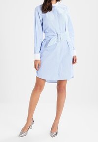 Hellblau gestreiftes Hemdkleid mit tailliertem Gürtel, langen Ärmeln und weißen Manschetten. Getragen mit silbernen High Heels.