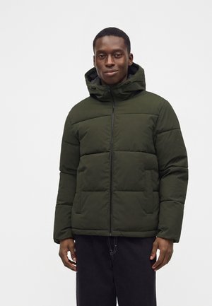 Man wearing a dark green hooded puffer jacket with front pockets and black pants, standing against a plain light background.