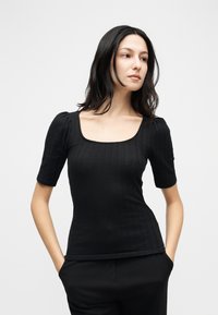 Γυναίκα με ίσια μαύρα μαλλιά, που φορά μια εφαρμοστή μαύρη ριμπ μπλούζα με κοντά φουσκωτά μανίκια και μαύρο παντελόνι, στέκεται με τα χέρια στις τσέπες.