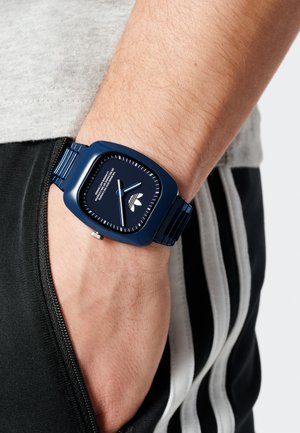 Orologio quadrato blu con cinturino in metallo. Il quadrante presenta indicatori delle ore e lancette bianchi, con un logo posizionato nella parte inferiore. Indossato al polso.