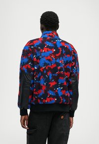 Fleece jacket with a multicolored red, blue, and black pattern; features black faux leather accents on sleeves and a high collar.