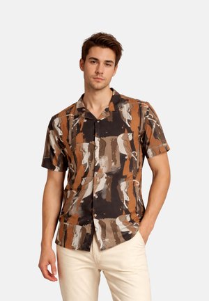Jeune homme portant une chemise à manches courtes avec un motif abstrait marron, noir et beige, debout avec une main dans la poche.