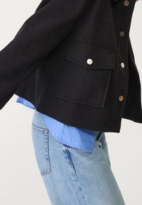 Osoba v světle modrých džínech, modré pruhované košili a černé bundě se stříbrnými patentkami a klopou na přední kapse.