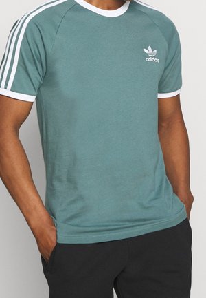 Homme portant un t-shirt Adidas bleu sarcelle avec un liseré blanc et trois bandes blanches sur les manches, les mains dans les poches d’un pantalon noir.