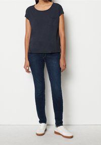 Persona che indossa una camicia a maniche corte blu scuro, jeans skinny blu scuro e sneaker bianche, in piedi contro un muro bianco.